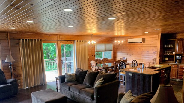 L’intérieur chaleureux d’un chalet avec un plafond en bois, un espace de vie comprenant un canapé et un pouf, une table à manger avec des chaises, et un coin cuisine.