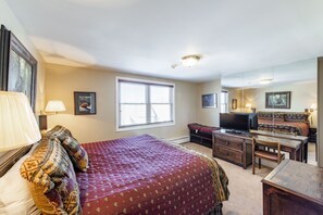 Une chambre avec un grand lit, une télévision, un bureau et une chaise. Il y a une fenêtre avec des stores, une lampe et un tableau encadré au mur.