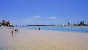 Ein Strand mit klarem Wasser, ein Steg im Hintergrund und Menschen, die am Ufer entlanggehen.
