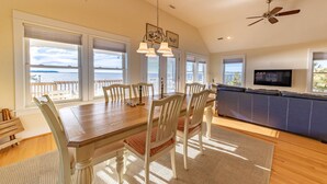 A dining area with a wooden table and white chairs, a ceiling fan, and a view of the ocean through large windows. - Sunset Joy - KDH MP 6, Soundfront, Sound View, Kayak, Community Boat Ramp, Pets OK, Flex Stays (Kill Devil Hills)