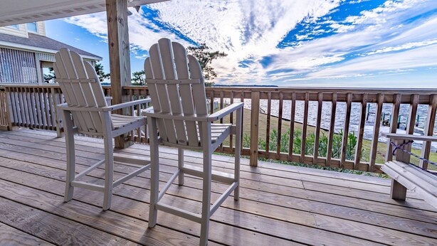 A wooden deck with two Adirondack chairs and a bench, overlooking a body of water with a wooden railing. - Sunset Joy - KDH MP 6, Soundfront, Sound View, Kayak, Community Boat Ramp, Pets OK, Flex Stays (Kill Devil Hills)