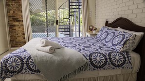 Een bed met een beddekleed met dessin, kussens, een opgerolde handdoek en een klein nachtkastje met een vaas en bloemen.