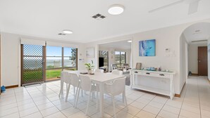 A bright, spacious living area with a dining table, white chairs, and a view of the outdoors through large sliding doors.