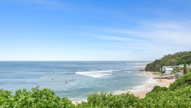A coastal view with a beach, ocean waves, and a hillside with buildings.
