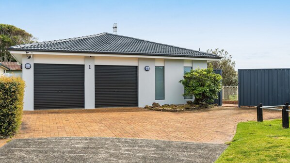 A modern two-story building with a tiled roof, two garage doors, and a small garden area with a bush.