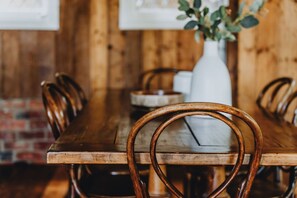 A wooden dining table with chairs, a white vase with greenery, and a brick wall in the background. - The Dairy., Perfectly positioned right in the heart of town located behind the majestic Green Gables (Castlemaine)