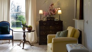 A room with a large window, a chandelier, a dresser, a blue armchair, a yellow armchair, and a small table with lamps and flowers.