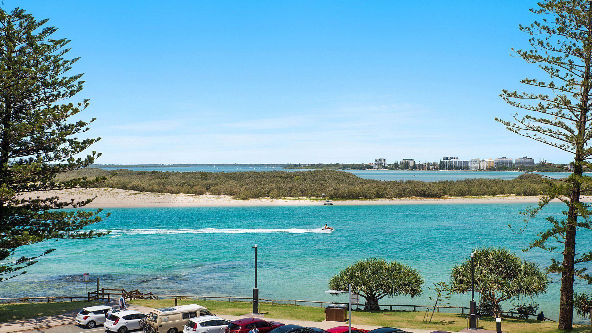 A coastal view with a beach, clear blue waters, and a distant cityscape.