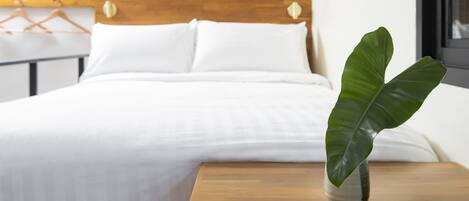 Una cama bien tendida con sábanas blancas y cabecera de madera. Una planta verde grande está colocada en una mesita de noche de madera al lado de la cama.
