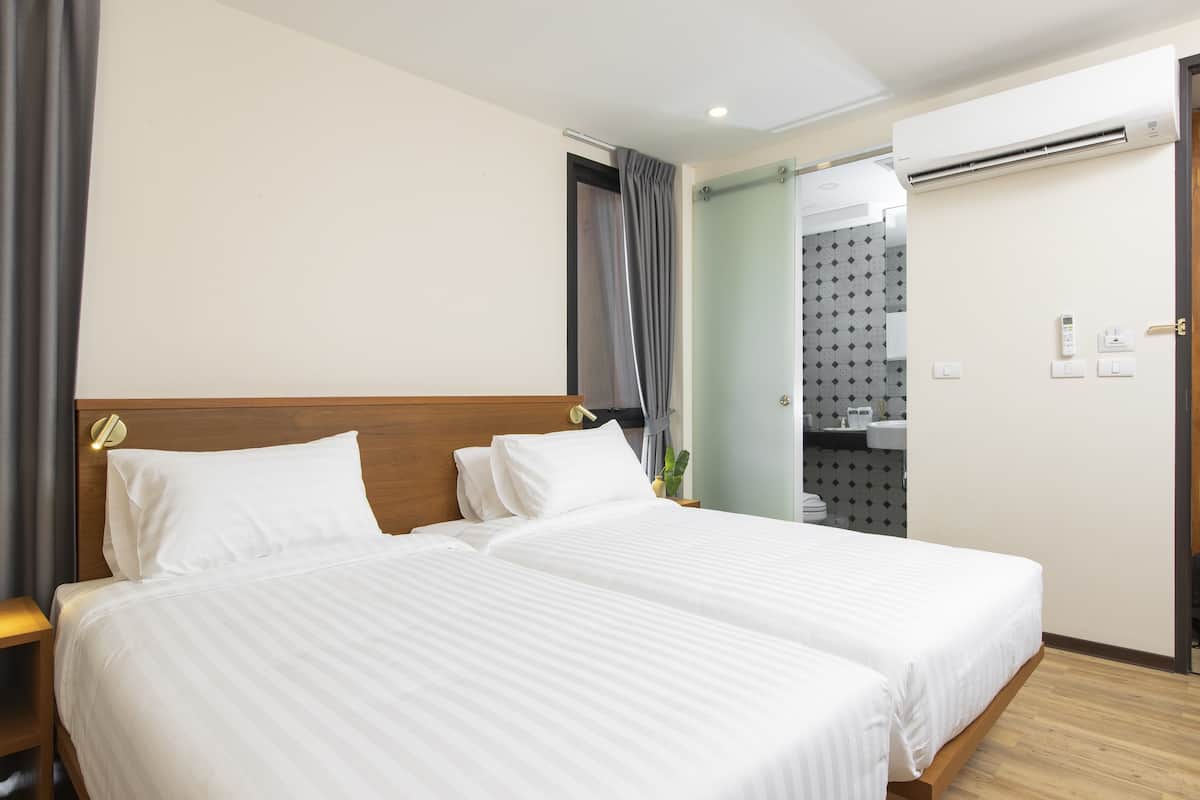 A hotel room with two beds, a wooden headboard, a bathroom with a shower, and a wall-mounted air conditioner.