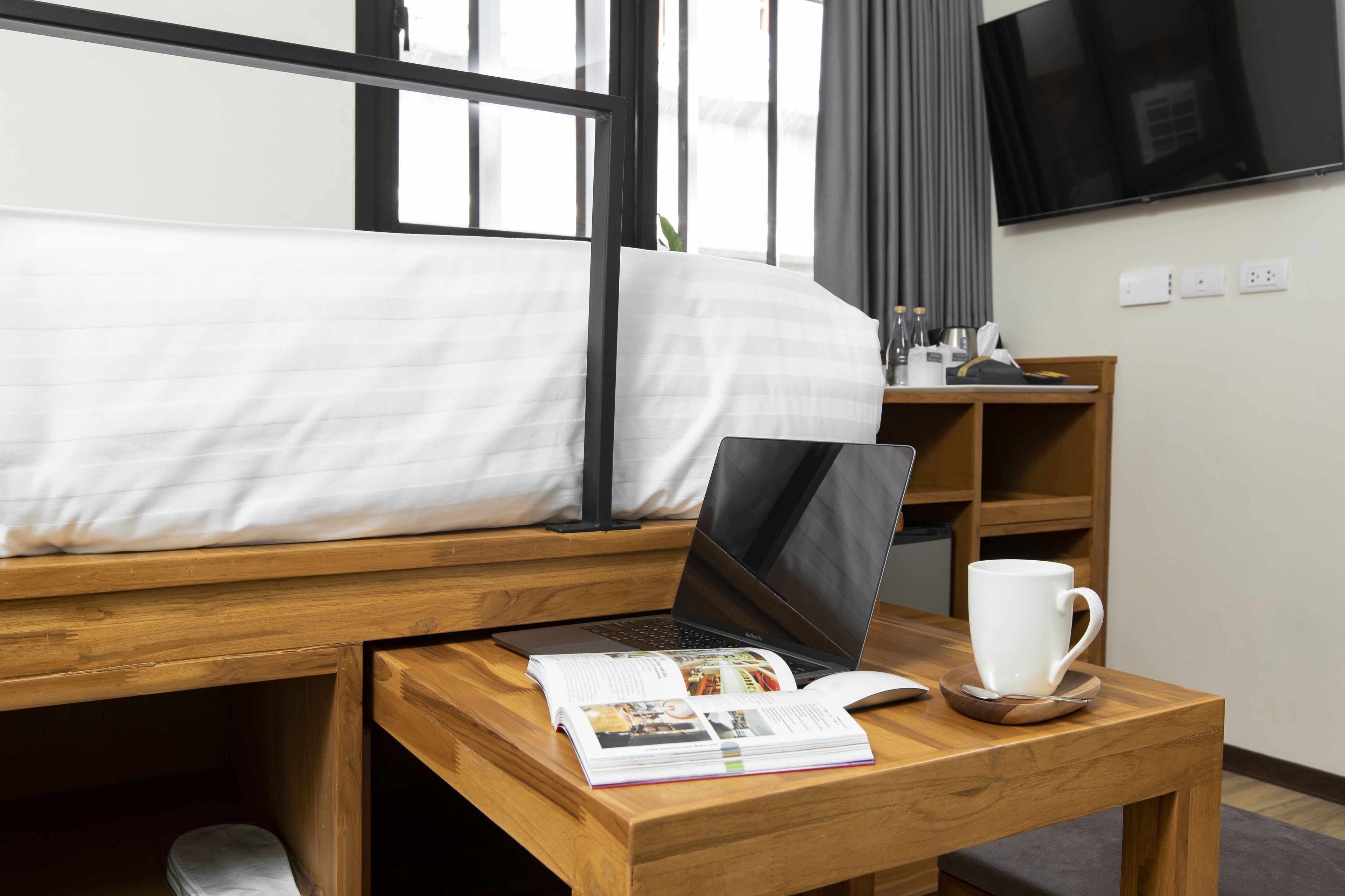 Un lit bien fait, avec un ordinateur portable et un magazine posés sur une table de chevet en bois, une tasse blanche et un téléviseur accroché au mur.