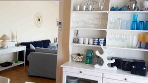 Un espacio de sala con un sofá, una mesa de centro y un televisor. Hay una estantería con diferentes vasos y vajilla.
