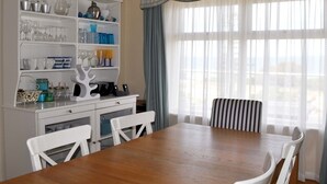 Un comedor con una mesa de madera y sillas blancas, un armario blanco con cristalería, y vistas a través de cortinas traslúcidas.
