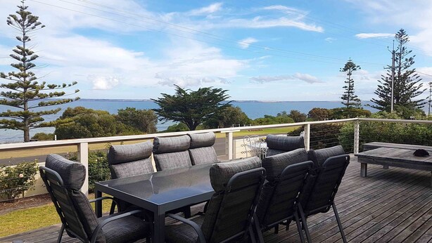 Una terraza al aire libre con una mesa y sillas, con vistas a un paisaje marino y arbolado.