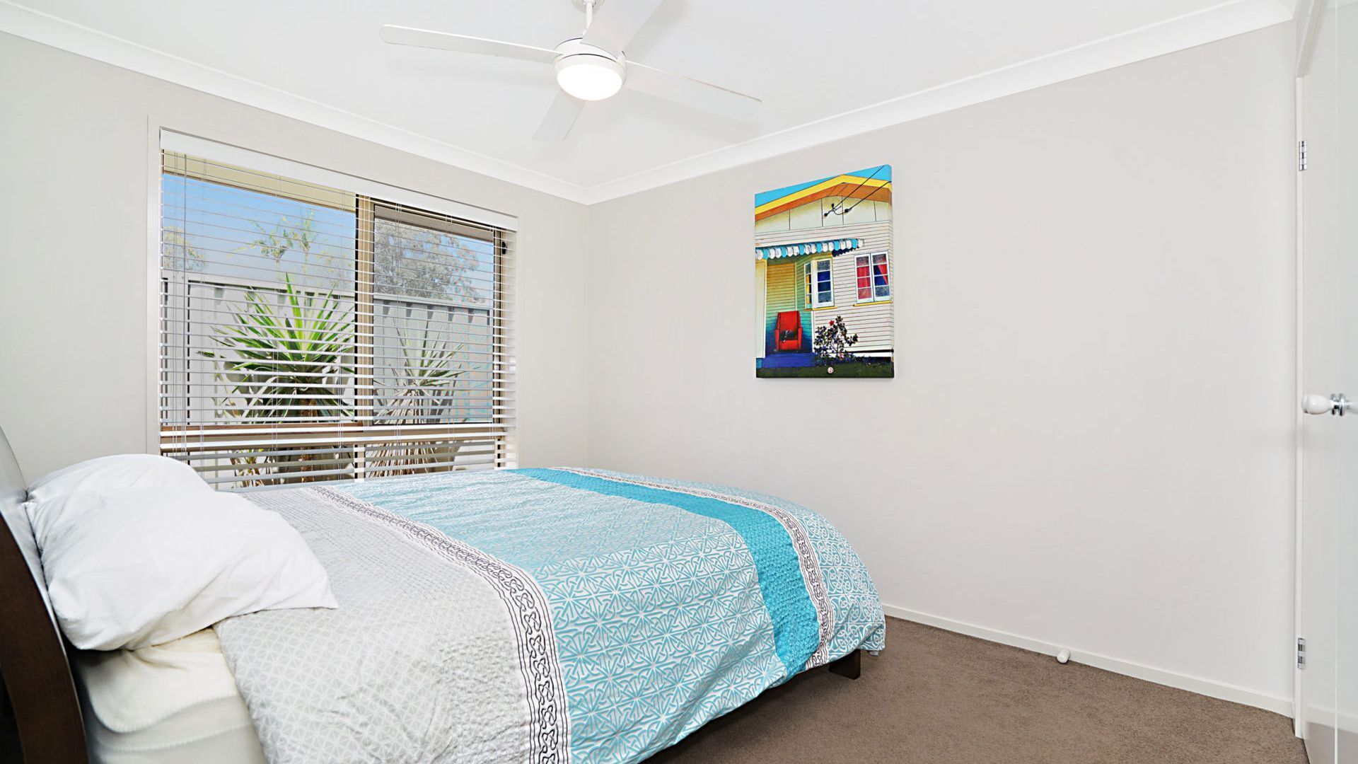 A bedroom with a bed, a ceiling fan, a window with blinds, and a wall-mounted picture.