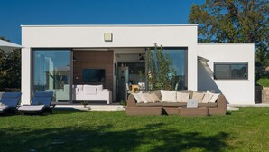 Een modern huis met een glazen entree, een woonkamer met een bank en een televisie, en een gazon met twee comfortabele stoelen.