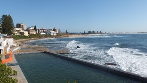 Vista costeira com um cais que se estende para o oceano, uma praia e edifícios residenciais ao fundo.