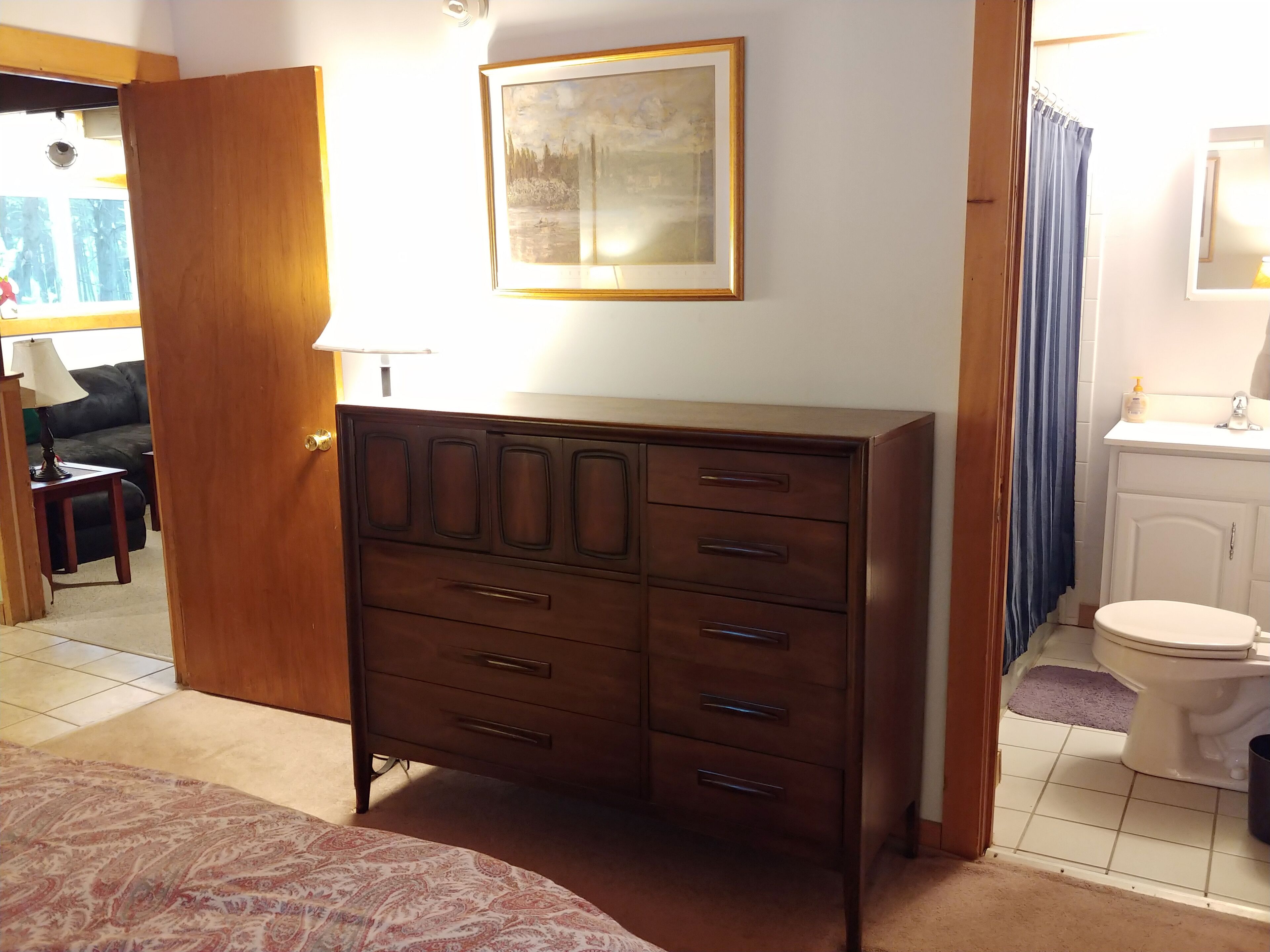 Un dormitorio con un aparador de madera, una cama con colcha estampada, un cuadro en la pared y un baño con inodoro y lavamanos.