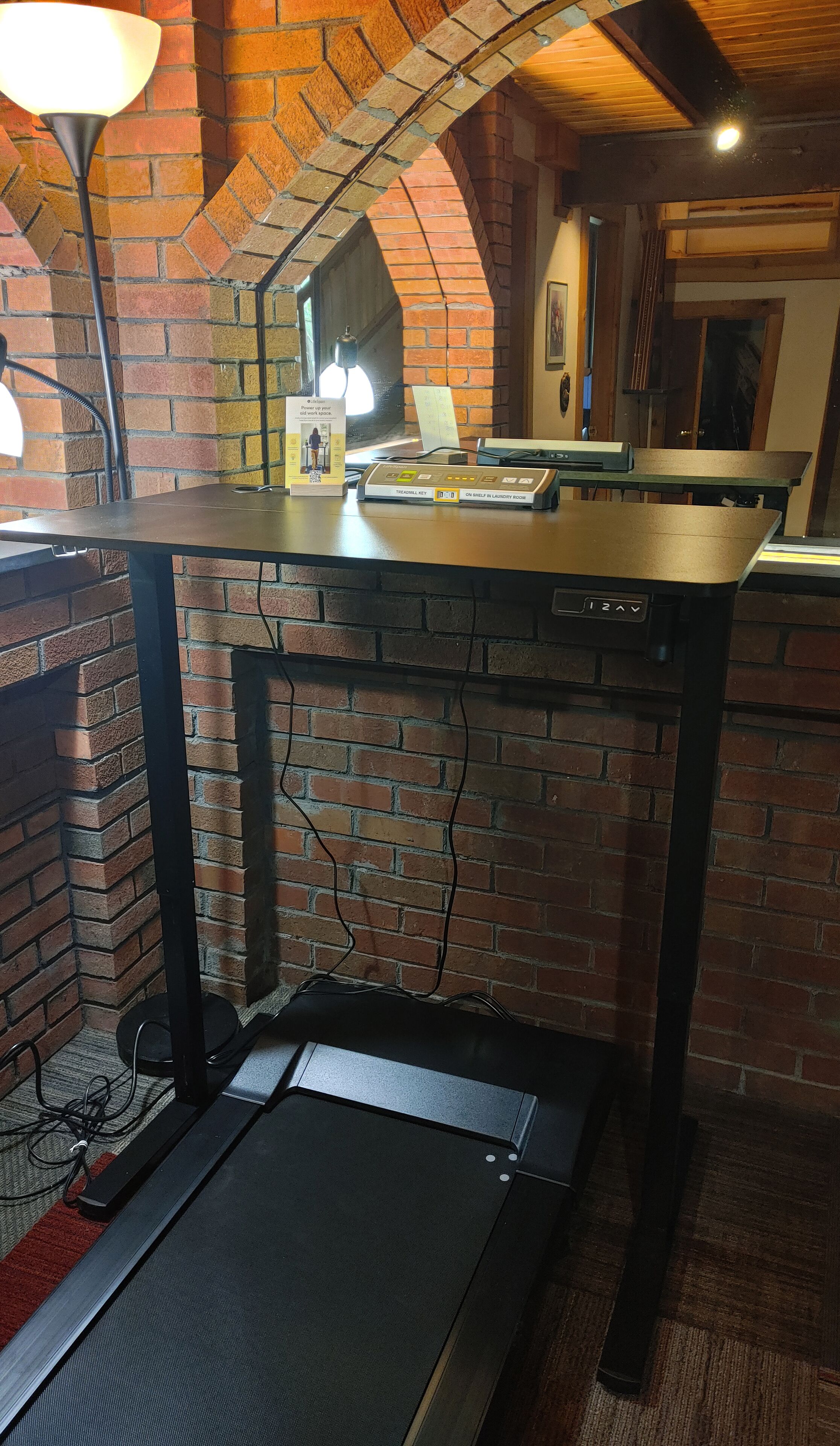 Et værelse med murstensvæg, løbebånd, skrivebord med computer og lampe.