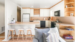 A compact kitchen with a central island, bar stools, a built-in microwave, and a wooden shelving unit.