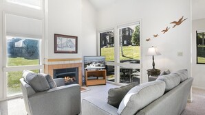 A modern living room with a fireplace, comfortable seating, a television, and large windows offering a view of the outdoors.