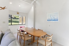 A dining area with a wooden table and chairs, a ceiling fan, a window with a view of a lawn, and a framed picture on the wall.