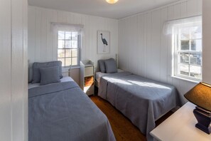 A room with two single beds, a small table, a lamp, a window, and a wall-mounted shelf with a framed picture.