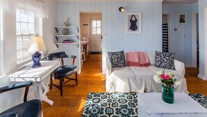 A cozy room with a white sofa, a small table with a vase of flowers, and a chair. There is a window with curtains, a lamp on a table, and a shelf with decorative items. - Across from Mayo beach walk to downtown! (Wellfleet)