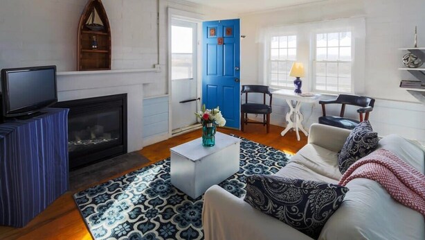 A living room with a fireplace, a blue door, a television, and a coffee table with a vase. - Across from Mayo beach walk to downtown! (Wellfleet)