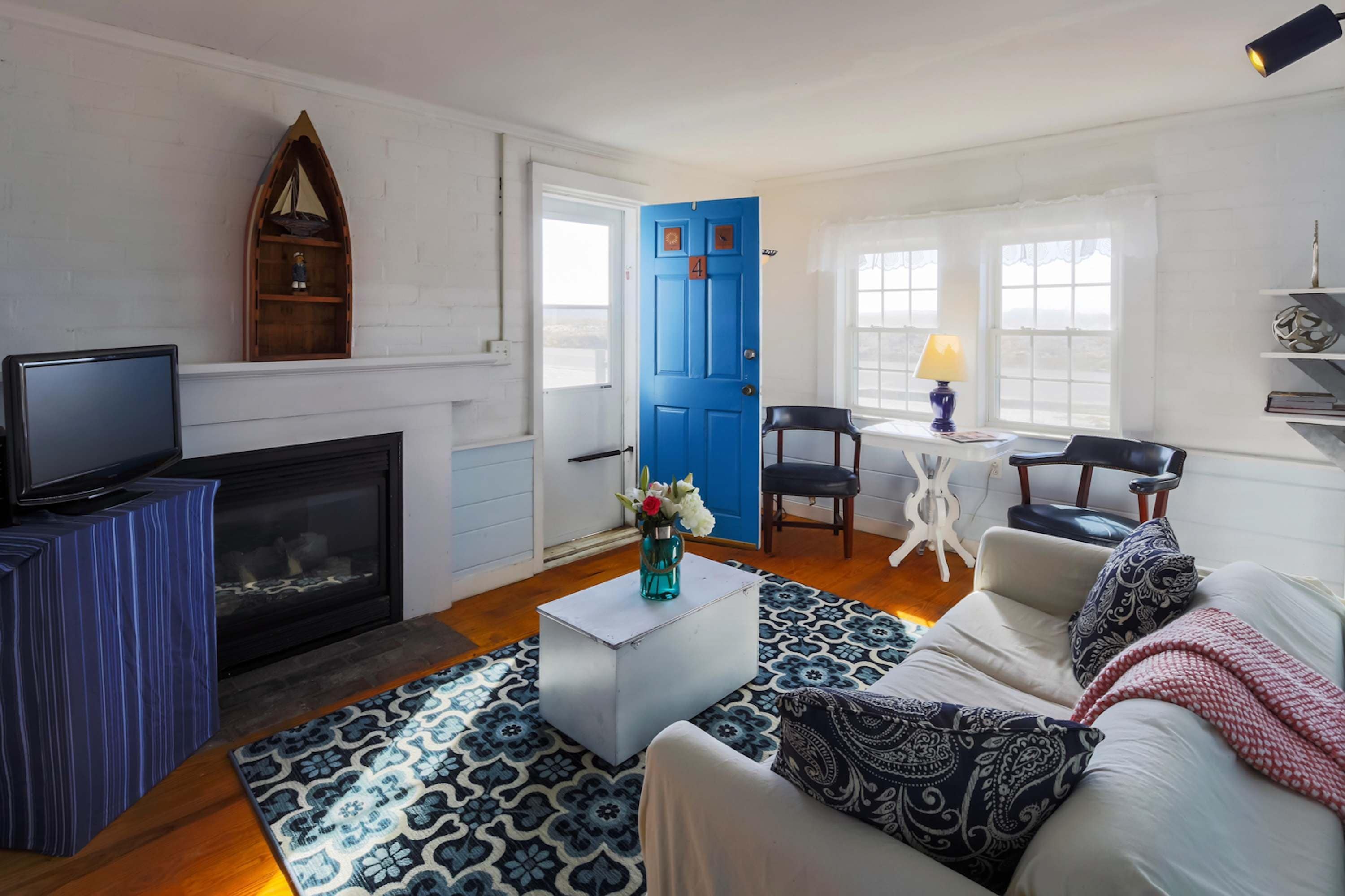 A living room with a fireplace, a television, a blue door, a dining area with chairs, and a sofa with patterned cushions.