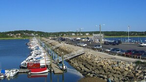 A marina with boats docked along a rocky breakwater, a parking area with cars, and a clear view of the water and distant shoreline. - Across from Mayo beach walk to downtown! (Wellfleet)