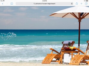 A beach scene with two wooden lounge chairs and a white umbrella, overlooking the ocean. - Walk To Beach-Relax-Private-Fun-Heated Pool-WiFi-Near All (Boynton Beach)