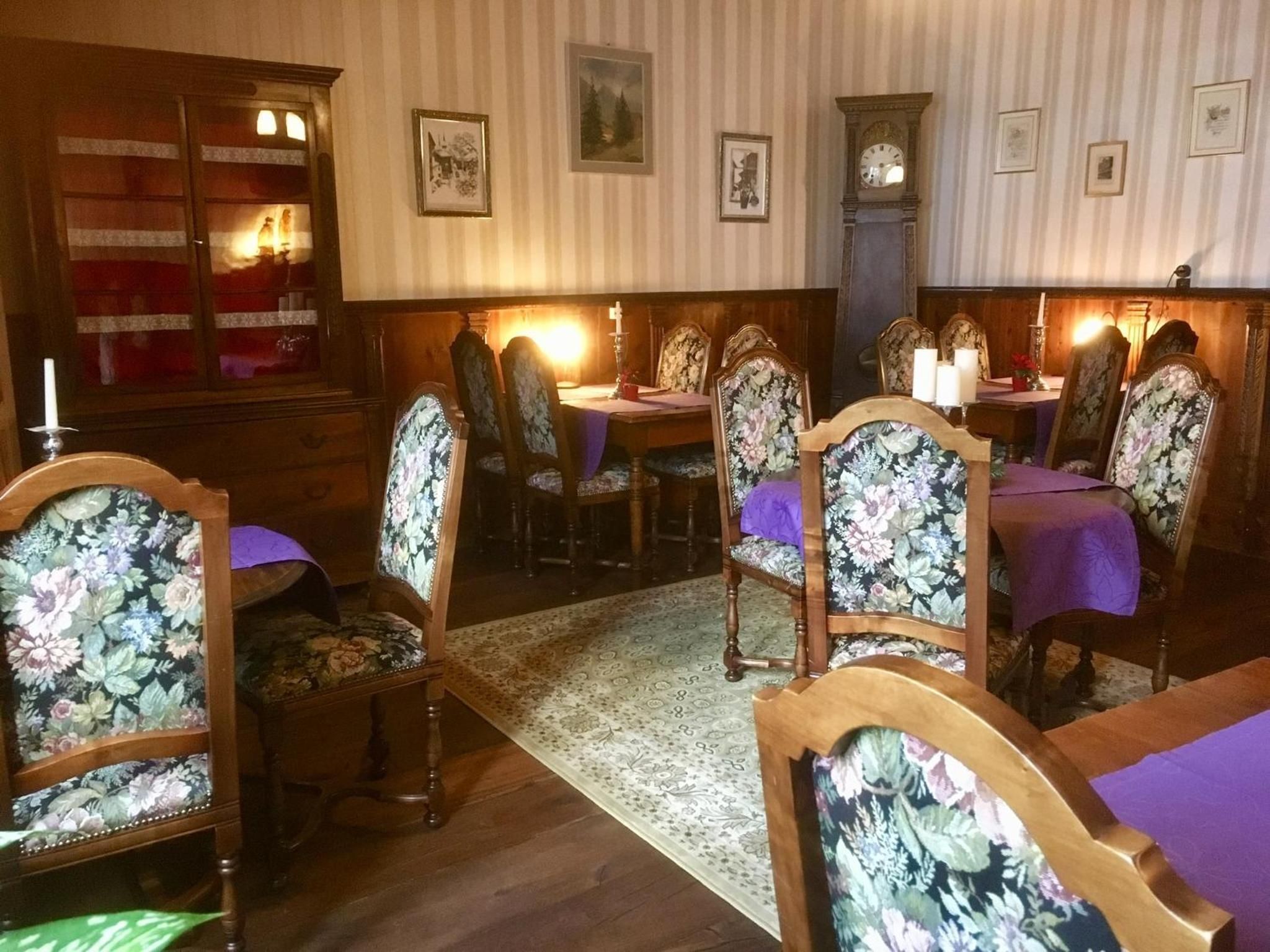 A dining area with wooden chairs and tables, a patterned rug, and framed pictures on the wall.