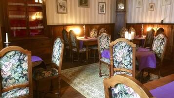 A dining area with wooden chairs and tables, a patterned rug, and framed pictures on the wall.