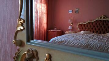 A room with a large mirror reflecting a bed with a tufted headboard, a nightstand with a lamp, and a window with curtains.