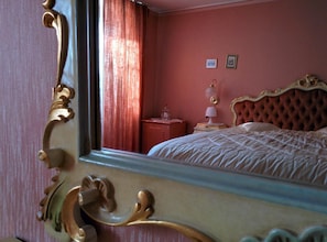 Une pièce dotée d’un grand miroir qui reflète un lit avec une tête de lit capitonnée, une table de chevet avec une lampe et une fenêtre avec des rideaux.