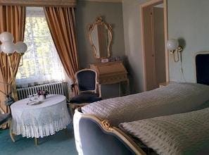 Une chambre à coucher avec un lit double, une coiffeuse avec un miroir, une chaise, une petite table ronde recouverte d’une nappe au crochet et un radiateur.