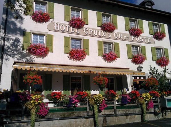 Un hotel con fachada blanca, contraventanas verdes y múltiples macetas con flores.