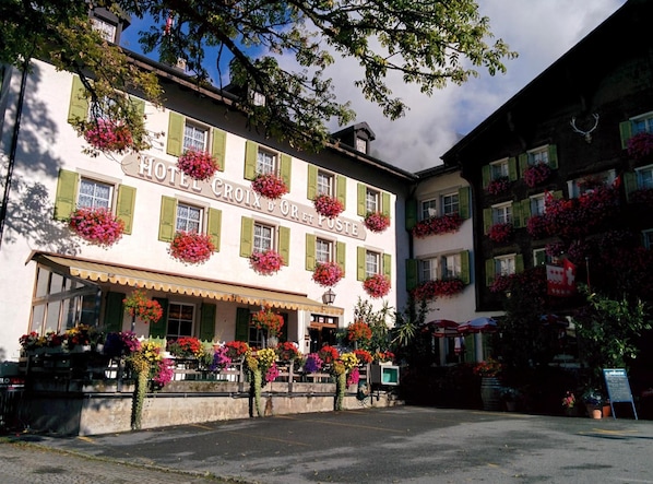 Un hôtel avec une enseigne indiquant « HÔTEL CROIX D’OR » et de nombreux jardinières fleuries sur les rebords des fenêtres.
