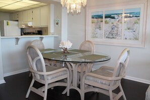 A dining area with a glass table surrounded by wicker chairs, a chandelier, and a beach-themed painting on the wall.