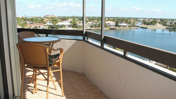 En balkon med fletmøbler, udsigt over en sø og omkringliggende boligbygninger.