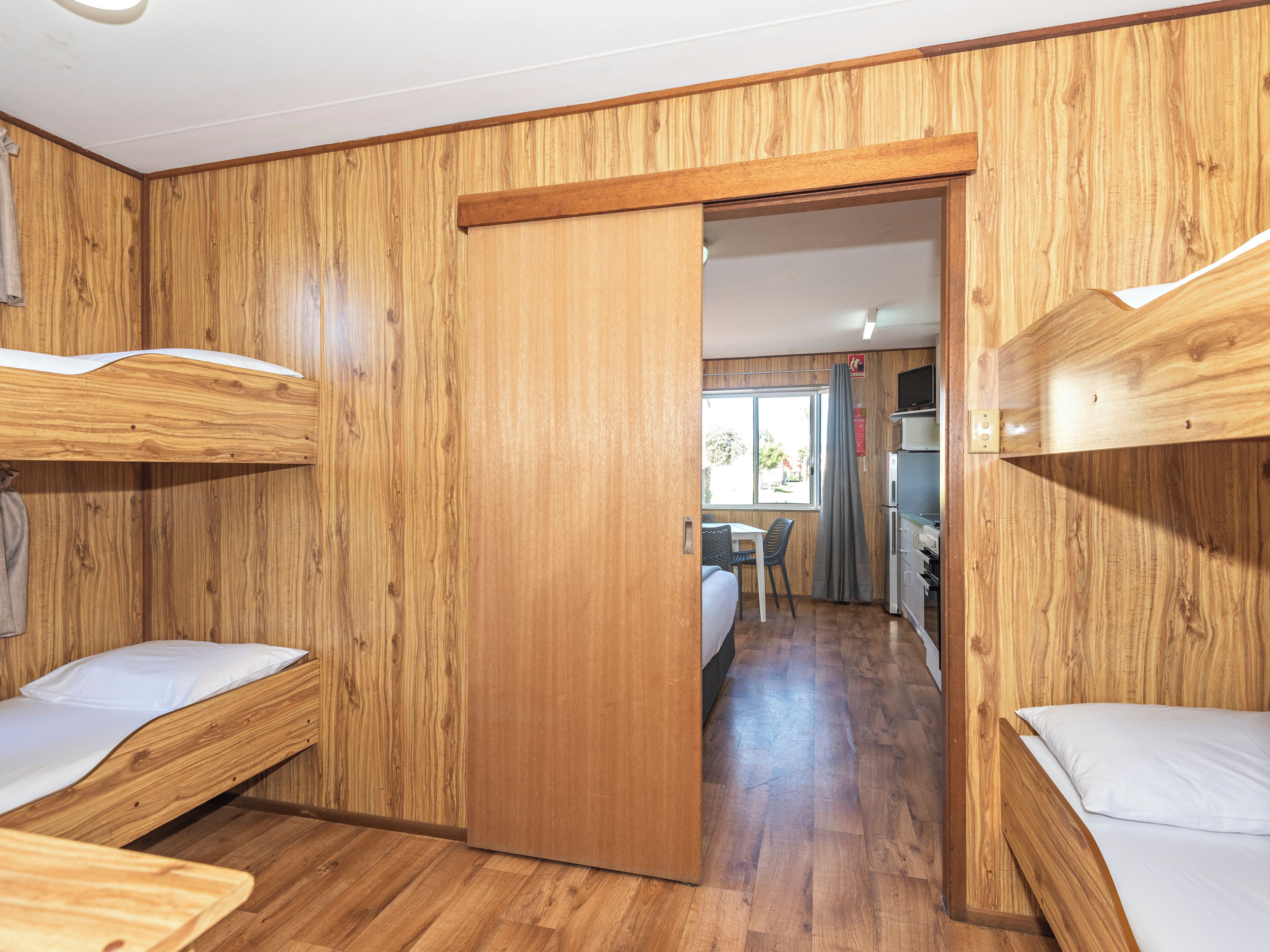 A room with wooden walls, bunk beds, a kitchenette, and a window with curtains.