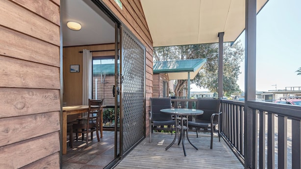 Une terrasse en bois avec une porte vitrée donnant sur une pièce, un espace extérieur aménagé avec des chaises et une table, et une vue sur les voitures garées et les bâtiments en arrière-plan.