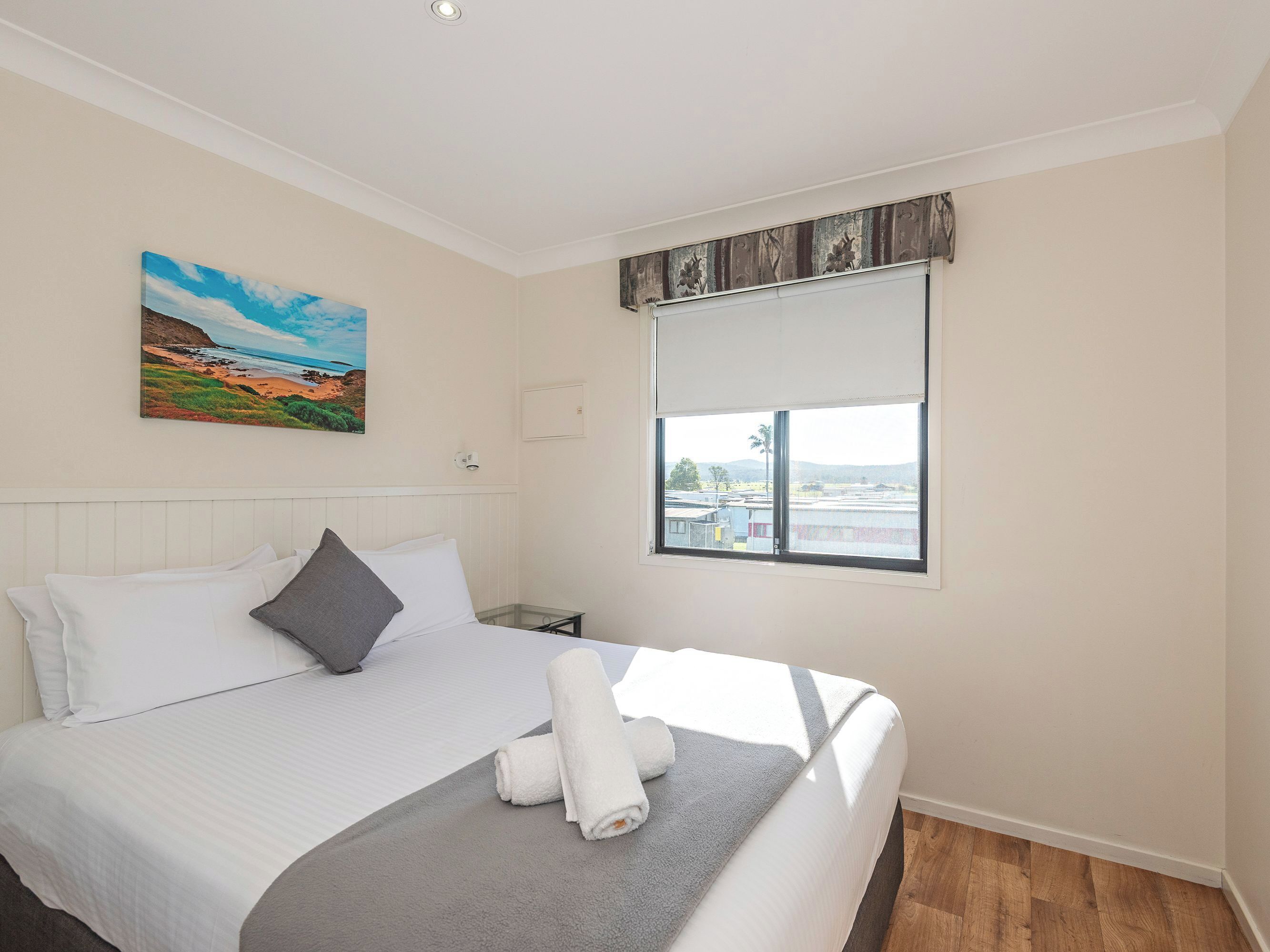 A neatly made bed with white linens and a grey blanket, a painting of a beach landscape on the wall, and a window with a view of buildings and hills.