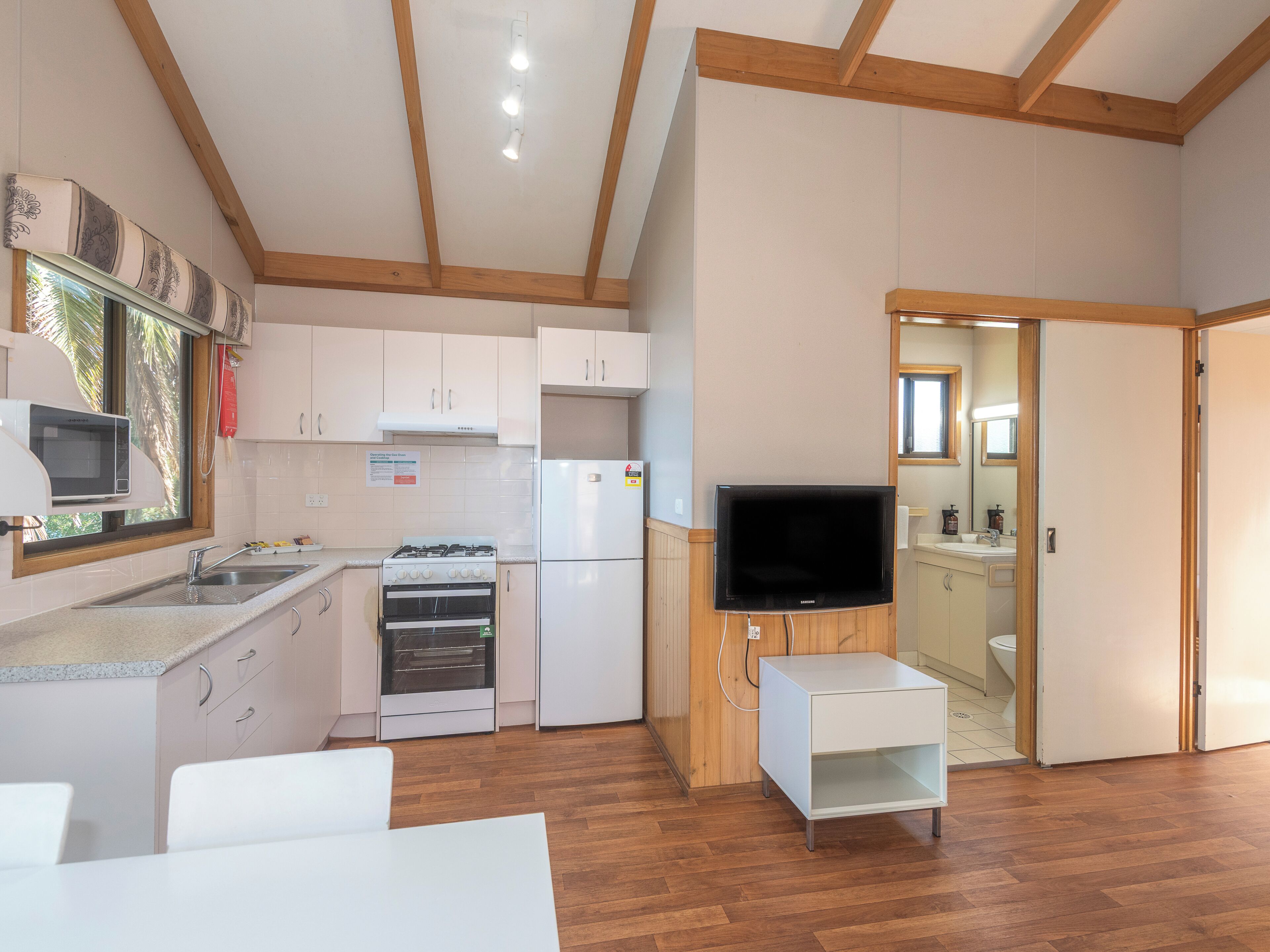 Eine kompakte Küche mit weißen Schränken, einer Mikrowelle, einem Herd und einem Kühlschrank. Ein Fernseher steht auf einem Holzboden und es gibt einen kleinen weißen Tisch.
