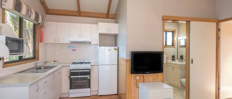 Eine kompakte Küche mit weißen Schränken, einer Mikrowelle, einem Herd und einem Kühlschrank. Ein Fernseher steht auf einem Holzboden und es gibt einen kleinen weißen Tisch.