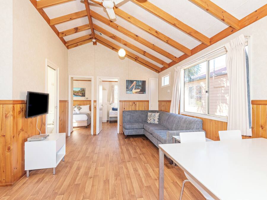 A spacious room with wooden beams, a flat-screen TV, a sofa, a dining table, and a window with curtains.
