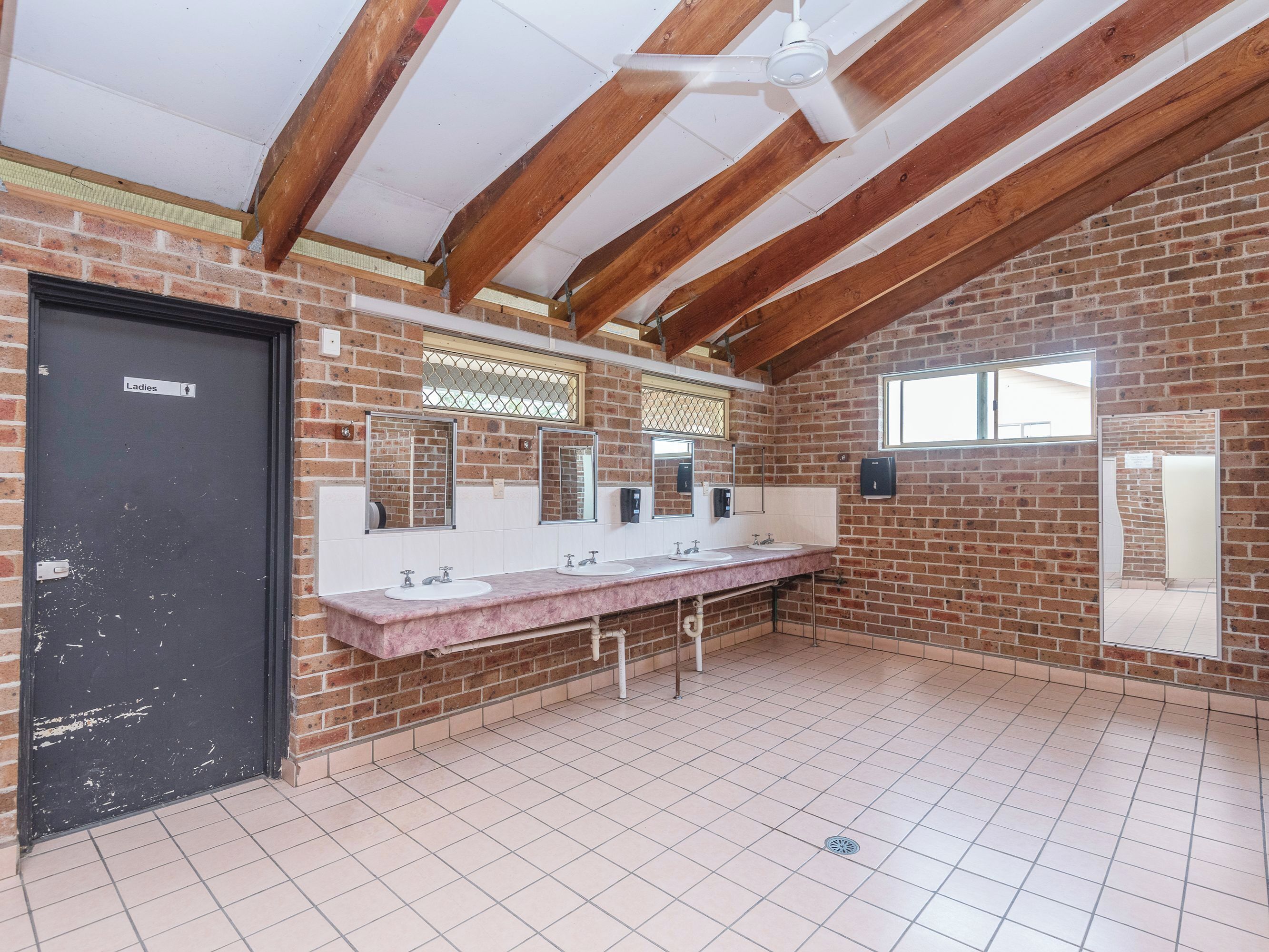 A restroom with a brick wall, a black door labeled 'Ladies,' a white ceiling fan, and a long vanity with three sinks.