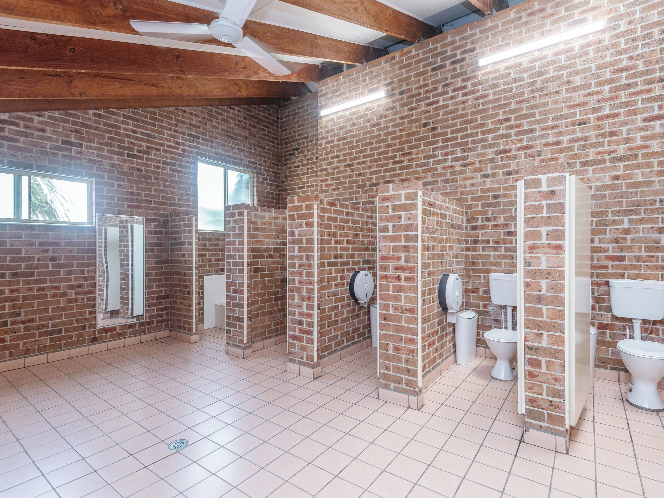Baño público con paredes de ladrillo, varios cubículos y un ventilador de techo.