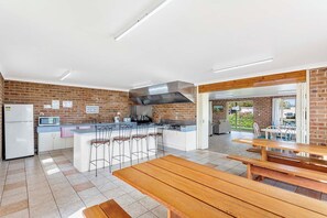 A modern kitchen with a central island, bar stools, a dining area with wooden tables, and a view of the outdoors. - Moruya Holiday Park (Moruya)